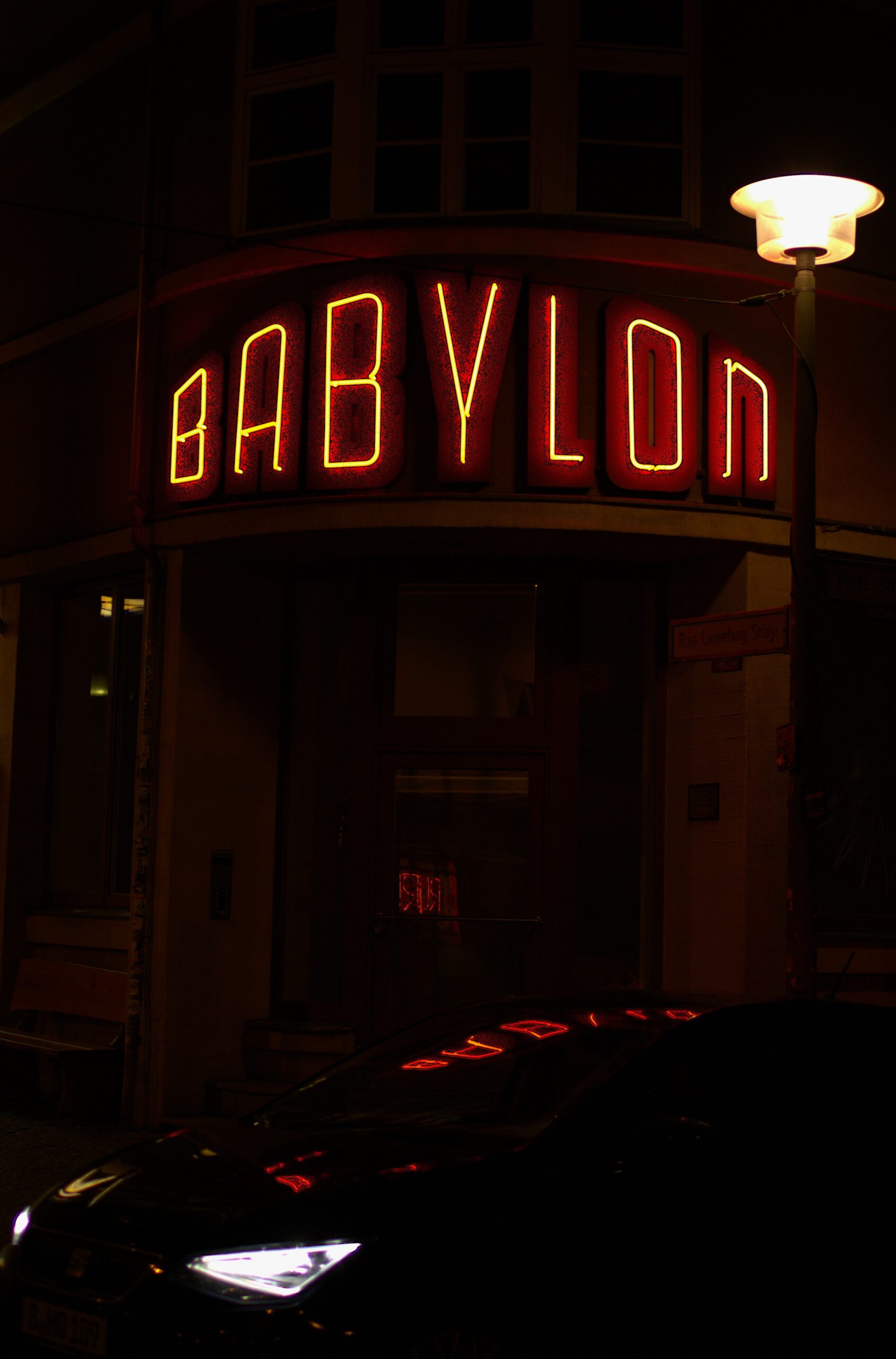 Red Babylon cinema sign above a dark street, with neon reflected on a car.