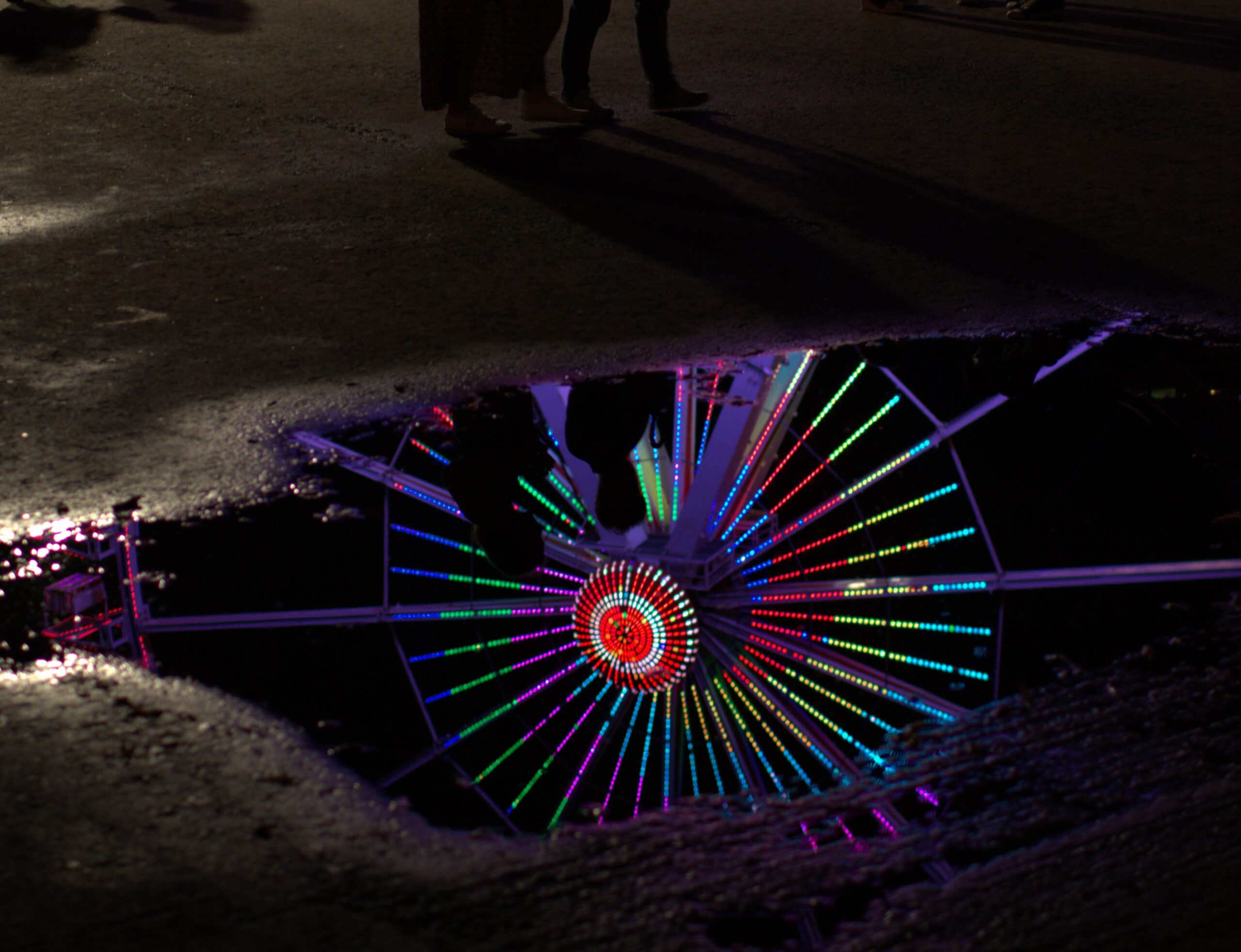 A couple beside a puddle reflecting colourful Ferris wheel lights and their silhouettes.