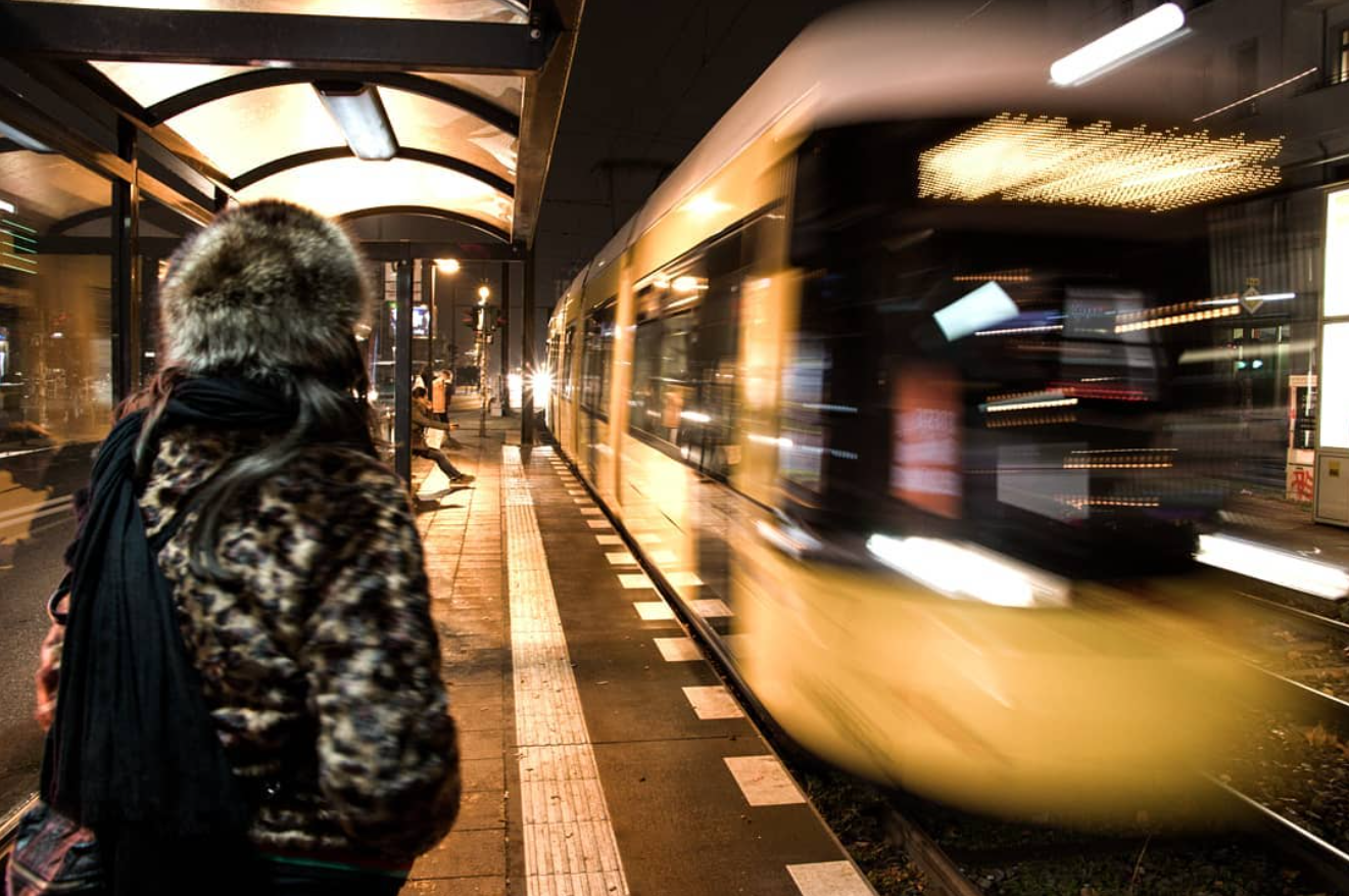 A woman waits beside a lit tram shelter as a yellow tram passes with motion blur.