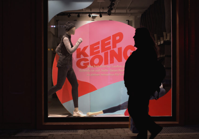 A silhouetted passer-by crosses an illuminated mannequin display with the words KEEP GOING.