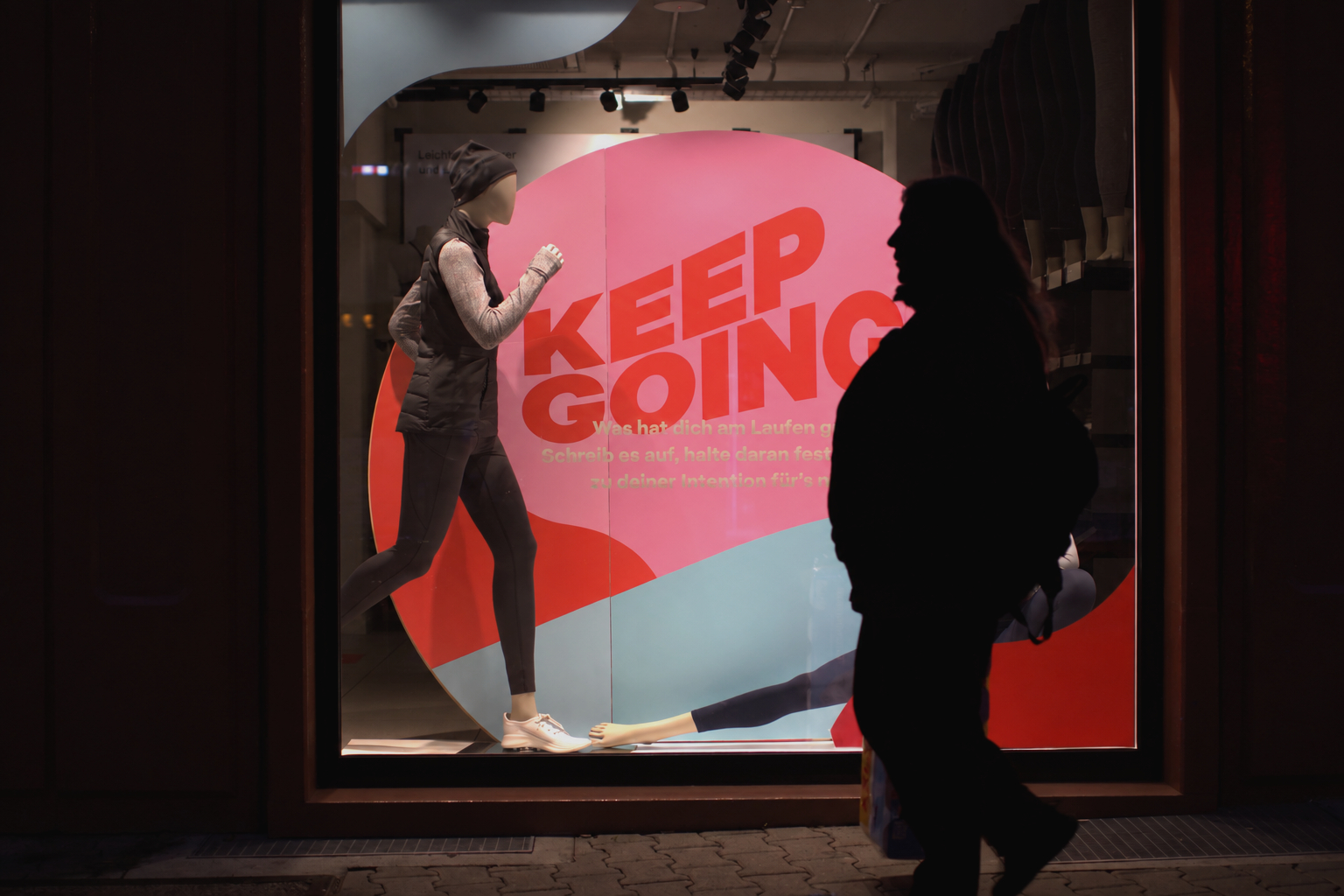 A silhouetted passer-by crosses an illuminated mannequin display with the words KEEP GOING.